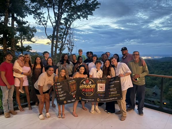 Cheerful multi-generational group photo on a mountain-overlook deck at sunset, holding a colorful promotional banner with rolling hills and a dramatic twilight sky in the background.