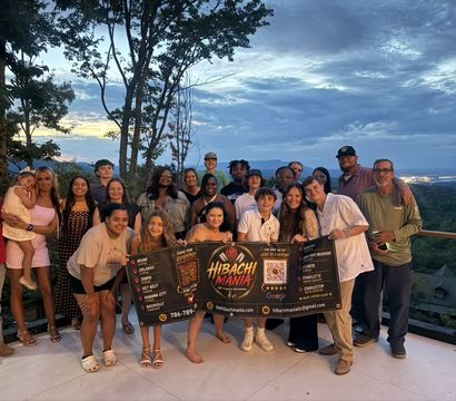 Cheerful multi-generational group photo on a mountain-overlook deck at sunset, holding a colorful promotional banner with rolling hills and a dramatic twilight sky in the background.