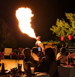 Fire-breathing performer shoots a dramatic flame at an outdoor dinner patio while silhouetted guests watch under red lanterns.