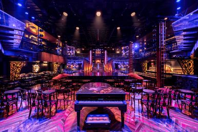 Vibrant cabaret-style live music venue interior with a grand piano in the foreground, round tables and chairs, two-tier balconies and a central lit stage under purple-blue spotlights