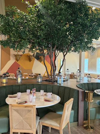 Modern restaurant interior with a large indoor tree growing from a central planter, green curved banquette seating around a white round table set with glasses and napkins, lantern candles on the planter ledge, wooden chairs, and a colorful geometric wall with decorative bull head mounts.