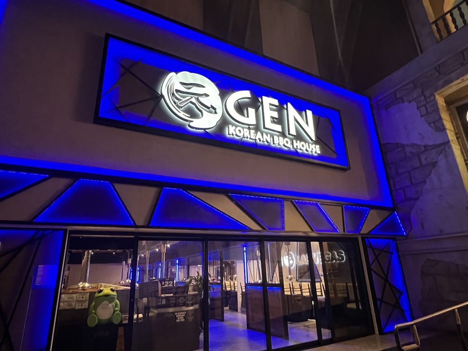 Nighttime exterior of a Korean BBQ restaurant with vivid blue LED trim and an illuminated circular logo above glass doors; interior booths and a stuffed frog visible through the window