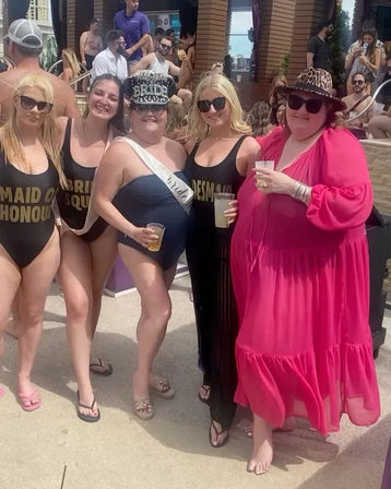 Bachelorette pool party at a resort — bride wearing a sash and sparkly BRIDE cap with bridesmaids in matching black swimsuits and a friend in a bright pink dress, all smiling and holding drinks.