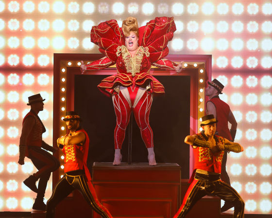 Extravagant drag performer in a red sequined sculptural costume with oversized shoulders and a showy updo, standing on a lit stage platform surrounded by four ringmaster-style dancers in red-and-black outfits and hats, bright marquee lights and a theatrical backdrop — high-energy cabaret performance.