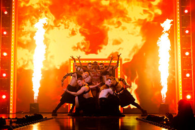 Show-stopping drag performer in a sparkling costume posed with backup dancers on a theater stage amid towering pyrotechnic flames and a fiery orange LED backdrop.