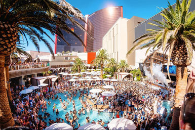 Daytime Las Vegas resort pool party with a huge crowd in and around the pool, palm trees, cabanas and umbrellas, DJ stage and high-rise hotel towers under a bright blue sky