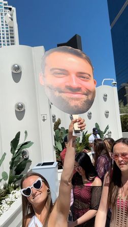 Sunny rooftop pool party in a city setting: people in sunglasses and summer outfits, one person holding a giant smiling face cutout on a stick, modern white poolside decor and tall buildings under a clear blue sky.