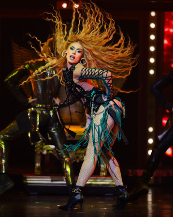 Energetic stage performer with long wavy blonde hair flying, dramatic makeup, hoop earrings and a green-and-black fringed costume with fishnet tights and high-heeled ankle boots, striking a dynamic dance pose under marquee lights with metallic-clad backup dancers.