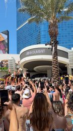 Daytime resort pool club party with a packed crowd holding up phones, bikini dancers on an elevated stage and a DJ booth beneath a palm tree against a glass high-rise backdrop.