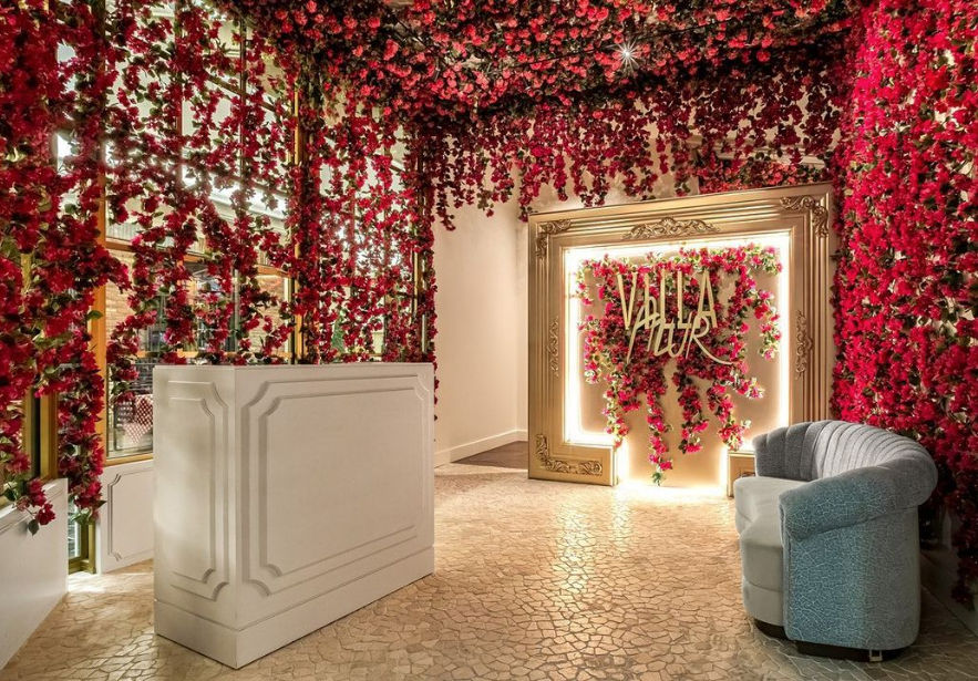 Floral-filled reception area with cascading red roses draping the walls and ceiling, a white reception desk, illuminated ornate gold frame with a flower-covered photo backdrop, and a curved gray lounge chair on a mosaic-tiled floor.