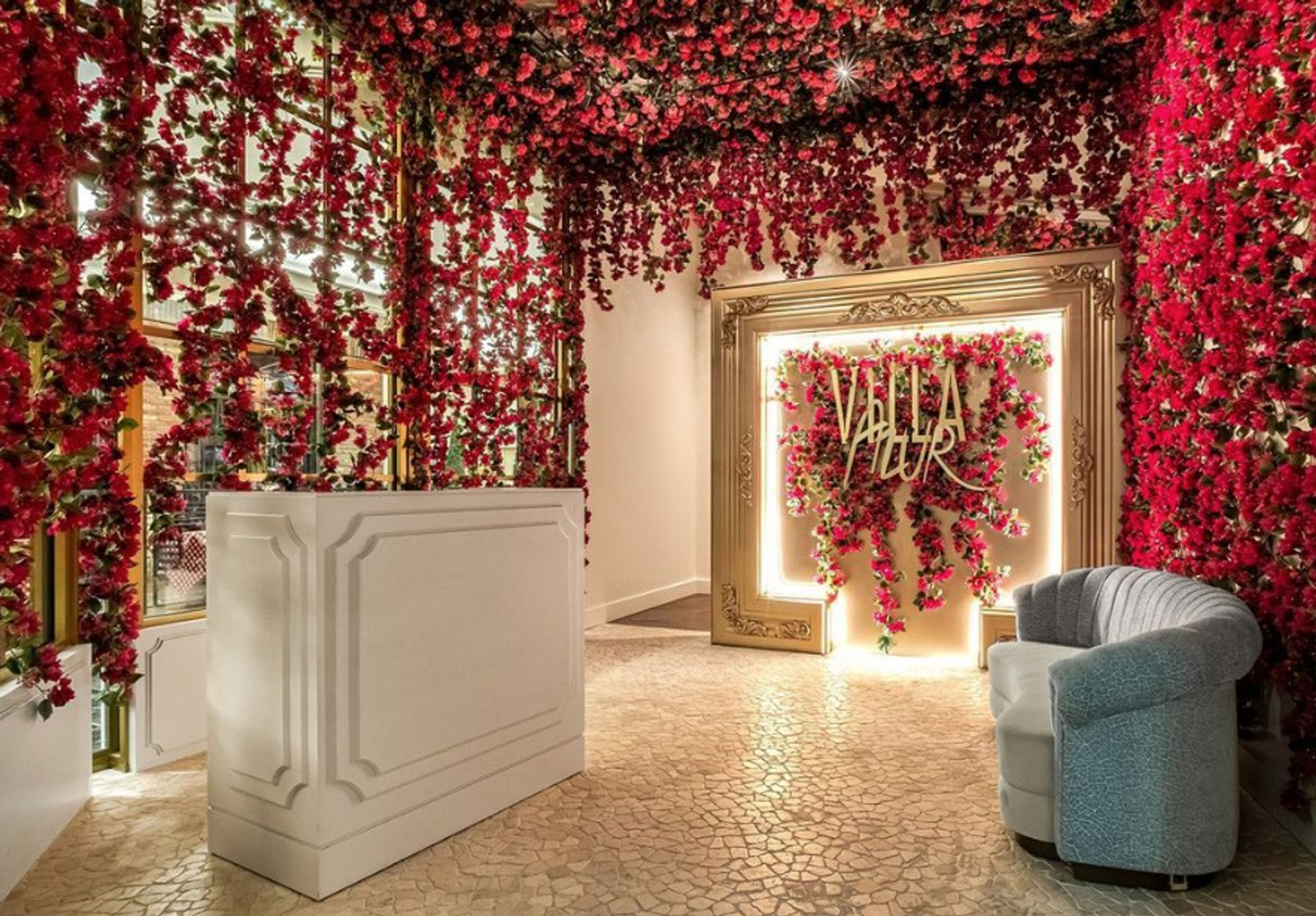 Floral-filled reception area with cascading red roses draping the walls and ceiling, a white reception desk, illuminated ornate gold frame with a flower-covered photo backdrop, and a curved gray lounge chair on a mosaic-tiled floor.