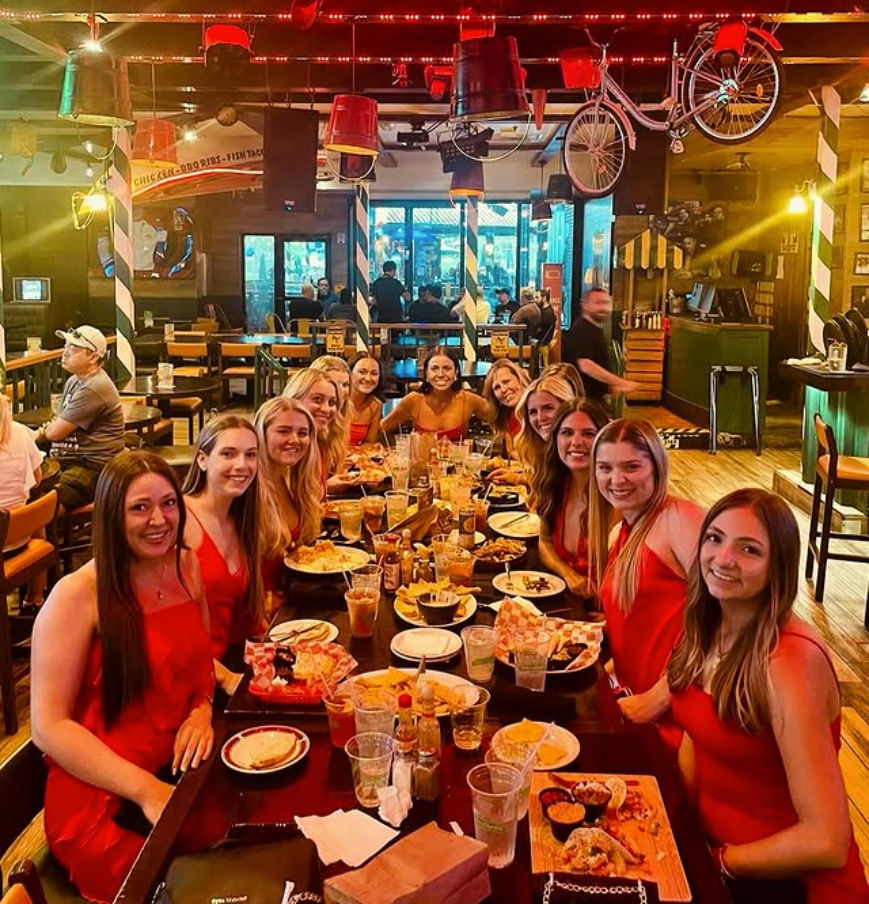 Smiling group of about a dozen women in red dresses gathered around a long wooden table at a lively indoor bar & grill, plates of appetizers and drinks on the table, warm string lights and quirky decor like a hanging bicycle and buckets.