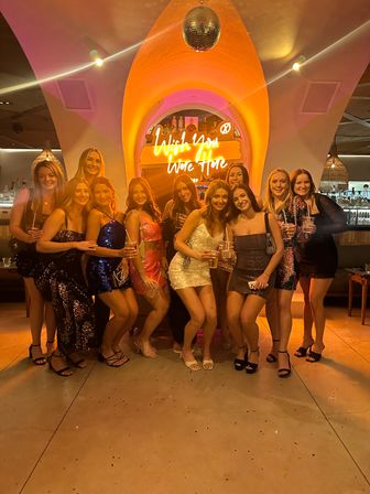 Group of friends in sparkly dresses posing with cocktails under a disco ball and neon “Wish You Were Here” sign in a warm-lit trendy bar interior.