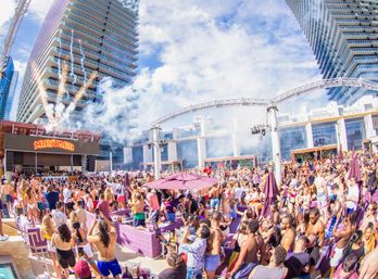 Vibrant daytime pool party at a luxury rooftop resort with a large crowd, DJ stage, fireworks, purple cabanas and surrounding high-rise buildings.