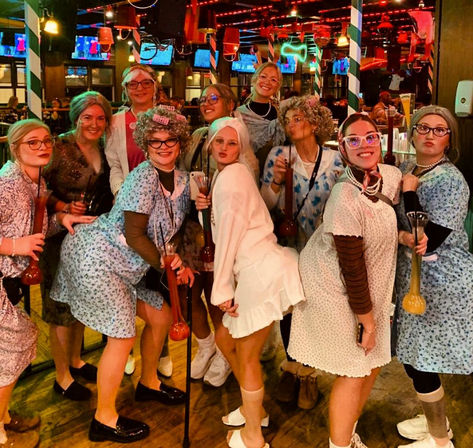 Group of women in playful 'elderly' costumes at a lively bar — curlers, glasses, shawls, canes and yard-long novelty drinks under neon lights