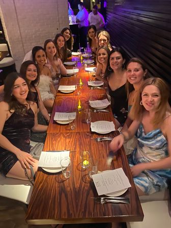 Group of women enjoying a candlelit girls' night out at an indoor restaurant, seated along a long wooden table with menus, glasses and small votive candles.