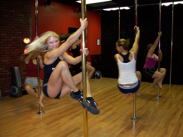 Group of women in a pole fitness class practicing spins on brass poles in a wood‑floored dance studio