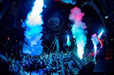 Packed nightclub dance floor with DJ at a lit booth pumping music as blue and pink smoke jets plume over a cheering crowd