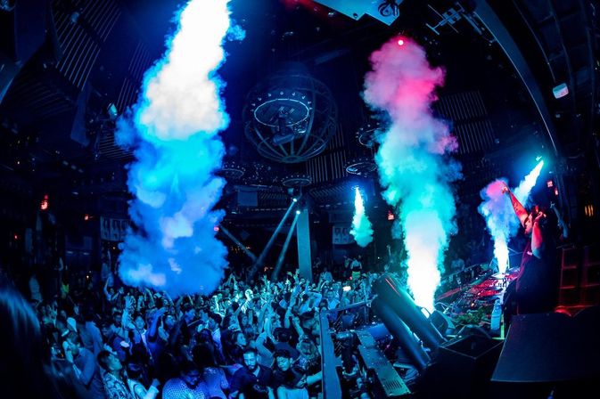 Packed nightclub dance floor with DJ at a lit booth pumping music as blue and pink smoke jets plume over a cheering crowd