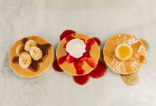 Top-down photo of three breakfast pancakes on a light surface: banana slices with chocolate sauce, strawberry-topped pancake with whipped cream and red syrup, and a butter-and-powdered-sugar pancake with maple syrup.