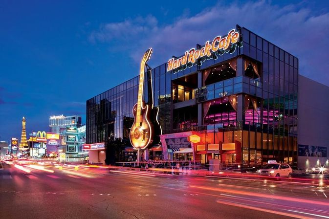 Dusk on the Las Vegas Strip — glass-front music-themed restaurant with a giant glowing guitar, colorful neon signs and streaking car light trails.