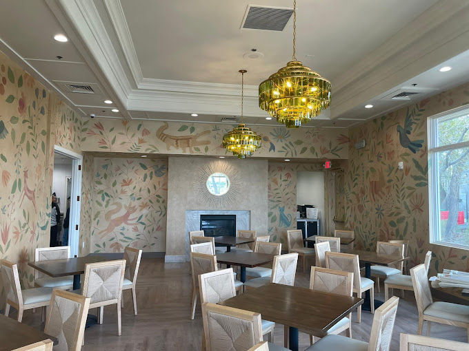 Sunlit indoor dining room with wooden tables and light-upholstered chairs, whimsical botanical and animal wall mural, gold tiered chandeliers, and a central fireplace.