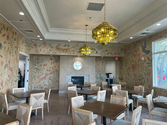 Sunlit indoor dining room with wooden tables and light-upholstered chairs, whimsical botanical and animal wall mural, gold tiered chandeliers, and a central fireplace.