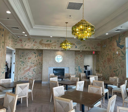 Sunlit indoor dining room with wooden tables and light-upholstered chairs, whimsical botanical and animal wall mural, gold tiered chandeliers, and a central fireplace.