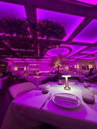 Upscale fine-dining restaurant interior bathed in magenta-purple LED light, white-tablecloth place setting with plates, napkin, glasses and small table lamp, floral ceiling installations and illuminated tree creating a romantic modern ambiance.