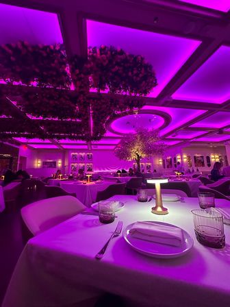 Upscale fine-dining restaurant interior bathed in magenta-purple LED light, white-tablecloth place setting with plates, napkin, glasses and small table lamp, floral ceiling installations and illuminated tree creating a romantic modern ambiance.