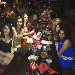 Dressed-up friends at a long restaurant table for a girls' night out, with cocktails, wine glasses, red napkins and candlelit ambiance