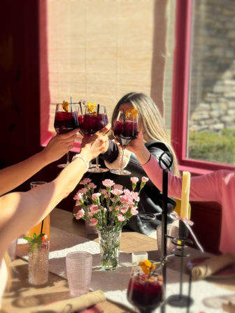 Sunlit café scene of four people clinking red sangria glasses over a table with pink carnations, candles, and brunch cocktails.
