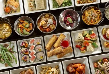 Top-down view of a colorful buffet of global small plates and appetizers — sushi rolls, crispy spring rolls with sweet chili dip, clams in broth, pasta, roasted vegetables and green beans on white dishes.