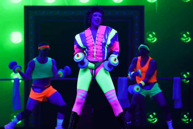 Performer in neon 80s-style aerobics costume lifting colorful dumbbells under blacklight on a theater stage with two backup dancers in fluorescent workout gear.
