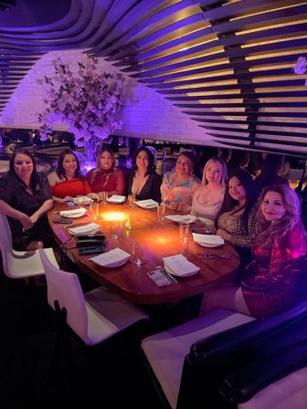 Eight women seated around a curved wooden table in a trendy, dimly lit restaurant booth with purple ambient lighting, large floral centerpiece, menus and candlelight — lively evening celebration.