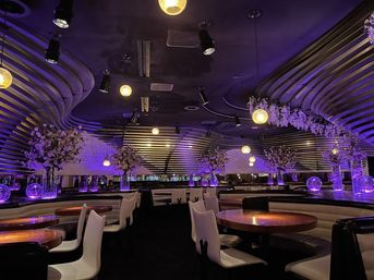 Upscale modern lounge interior with curved slatted ceiling, purple ambient lighting, hanging globe pendant lights, round wooden tables, white cushioned chairs, tall floral centerpieces in glass vases and a sleek bar in the background