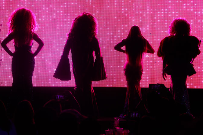 Silhouetted drag performers in glittering gowns strike poses against a vibrant pink LED backdrop on a nightclub cabaret stage