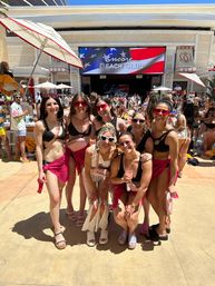 Seven friends in swimwear and pink sarongs posing at a crowded outdoor pool club day party, large LED screen with an American flag in the background and a sunny poolside vibe.
