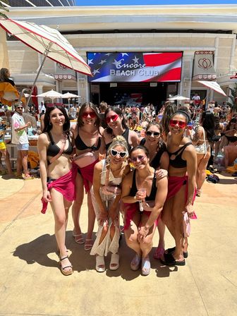 Seven friends in swimwear and pink sarongs posing at a crowded outdoor pool club day party, large LED screen with an American flag in the background and a sunny poolside vibe.