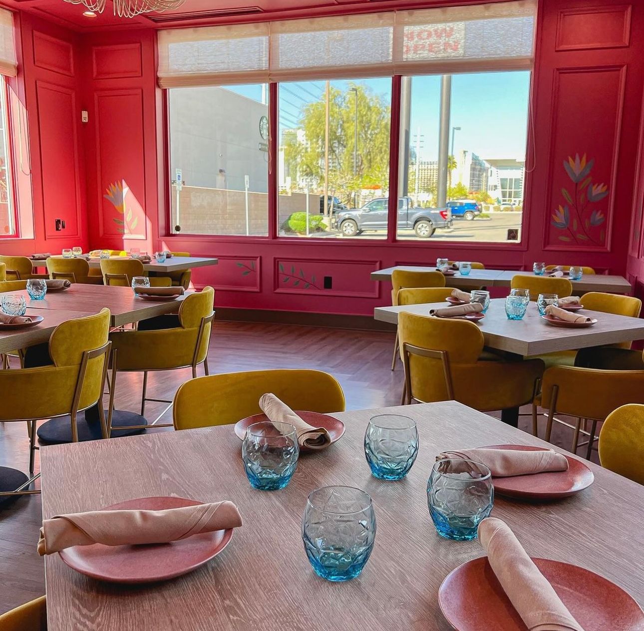 Bright pink restaurant interior with mustard-yellow chairs and light wood tables set with coral plates, beige napkins and textured blue water glasses, large windows offering an urban street view with parked cars