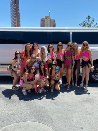 Bachelorette party group of women in bright pink outfits posing in front of a white stretch limousine on a sunny city street with tall hotel towers in the background.