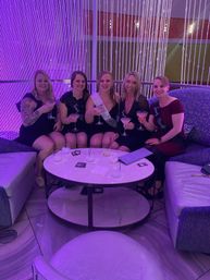 Five women in a purple-lit lounge seated on curved sofas with a beaded curtain backdrop, smiling and toasting with martini cocktails — playful bachelorette/girls’ night out celebration.