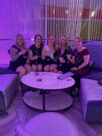 Five women in a purple-lit lounge seated on curved sofas with a beaded curtain backdrop, smiling and toasting with martini cocktails — playful bachelorette/girls’ night out celebration.
