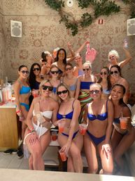 Group of friends in colorful bikinis at a sunny pool cabana bachelorette party, smiling and holding pink cocktails, sunglasses and playful props against a patterned wall with greenery.