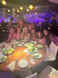 Smiling bride wearing a veil and friends pose around a round wooden table with salads and drinks during a bachelorette dinner in a dimly lit modern restaurant with purple accent lighting and hanging globe lamps.