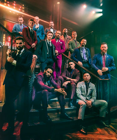 Group of men in colorful tailored suits posing on a wooden staircase inside a moody lounge with dramatic stage lighting — stylish, formal group portrait.