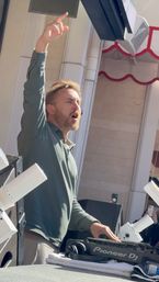 Outdoor daytime DJ set — bearded DJ in a green long-sleeve with one arm raised and the other on a DJ controller, surrounded by speakers and stage lights while fans record on smartphones.
