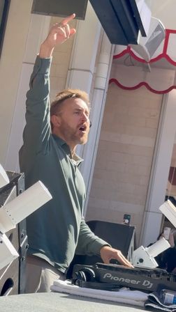 Outdoor daytime DJ set — bearded DJ in a green long-sleeve with one arm raised and the other on a DJ controller, surrounded by speakers and stage lights while fans record on smartphones.