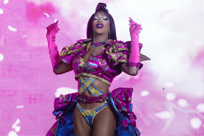 Drag performer in vibrant pink-and-blue embellished costume and long pink gloves, dramatic makeup, striking a pose on an LED-lit stage