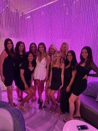 Group of women in cocktail dresses, one wearing a white mini dress, posing and smiling in front of a glowing purple beaded curtain at a trendy lounge — night out photo.